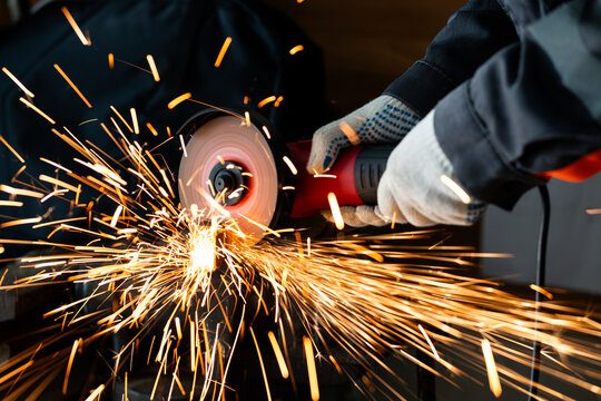Close-up of a worker in gloves cutting metal with an angle grinder tool in a workshop. Bright sparks flying from the rotating abrasive disc, industrial manufacturing process