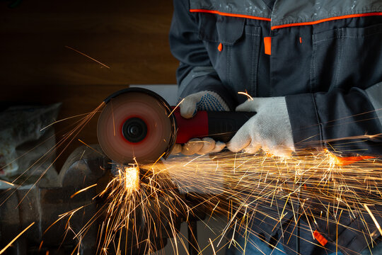 Close-up of a worker in gloves cutting metal with an angle grinder tool in a workshop. Bright sparks flying from the rotating abrasive disc, industrial manufacturing process.