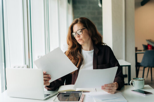 Woman reviewing documents at a laptop in a bright home office while working remotely and focused