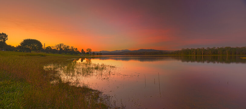 A wide panoramic sunset view over Lake Tinaroo in the Atherton Tablelands, Far North Queensland, with warm orange and purple evening light reflecting across calm water, distant hills, lakeside trees