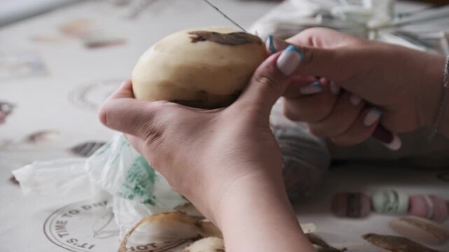 A close-up view of female hands carefully peeling the skin off a raw potato with a sharp paring knife at a kitchen table. Concept of homemade cooking, fresh vegetable preparation, and