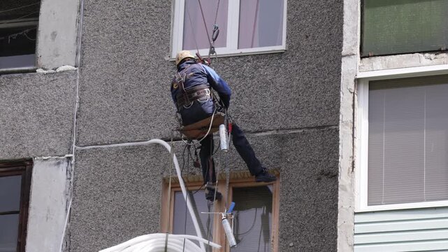 Worker suspended at height performs facade maintenance, applying insulation to seal gaps between concrete panels.