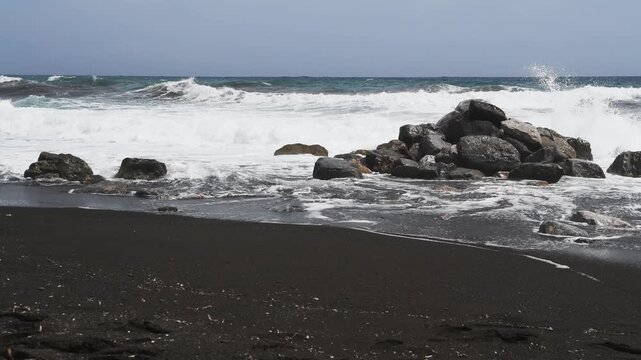 Waves on the beach in windy day. Perissa beach with black volcanic sand in Santorini island, Greece. Blue sea and the blue sky. 
