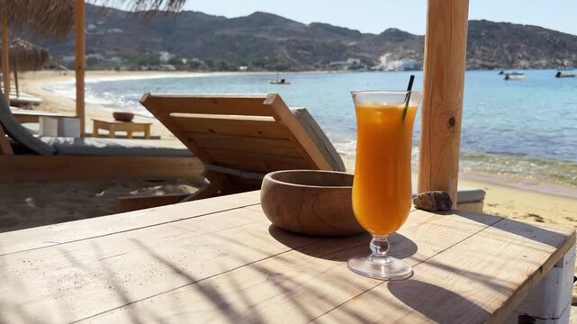 Beautiful beach in Ios island, Greece. A glass of fresh orange juice on the table. Chaise lounges with sun umbrellas on the beach. Blue sea and yellow sand. Travel and summer vacation concept. 
