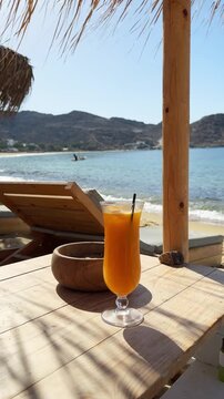 Beautiful beach in Ios island, Greece. A glass of fresh orange juice on the table. Chaise lounges with sun umbrellas on the beach. Blue sea and yellow sand. Vertical video