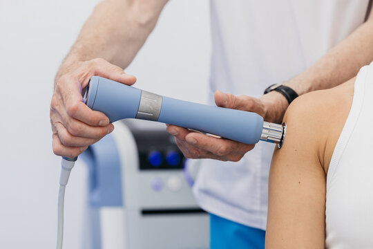 Doctor administering shockwave therapy to a patients shoulder using a handheld device in a clinical setting with medical equipment visible in the background
