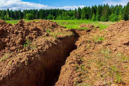 Freshly excavated trench in loamy soil across a rural field, showing raw earth piles with a lush green coniferous forest line in the background under a bright summer sky