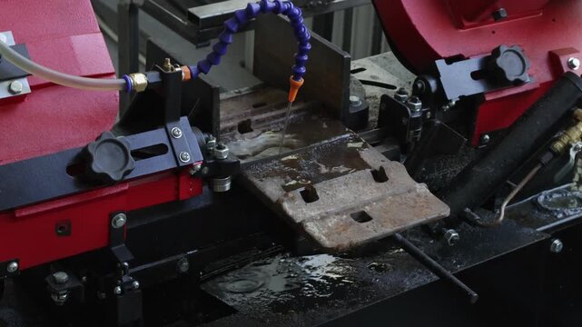 Industrial band saw cuts through a heavy metal plate. Coolant spray from flexible hose keeps blade sharp, creating dynamic mist.