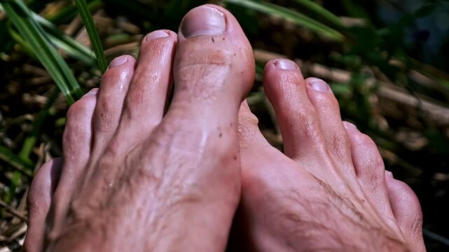 A macro shot captures male bare feet with visible hair and veins resting on the ground. The wiggling toes reveal details of skin texture and toenails in natural light.
