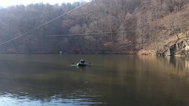 An overhead aerial drone view of a small inflatable boat with two people rowing across calm, dark lake water near a long, straight white safety buoy dividing line.

