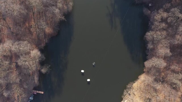 An overhead aerial drone view of a small inflatable boat with two people rowing across calm, dark lake water near a long, straight white safety buoy dividing line.

