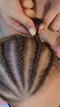 Female hands with yellow manicure braiding tight cornrows into brown hair in close up view. The concept represents skilled hairstyling and summer beauty care trends.