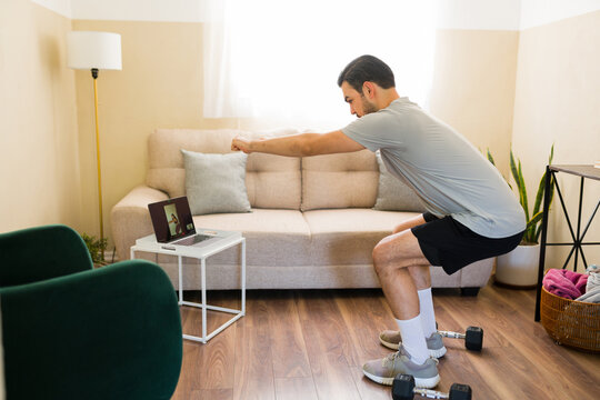 Latin man doing squats during virtual fitness class with online personal trainer on laptop at home gym