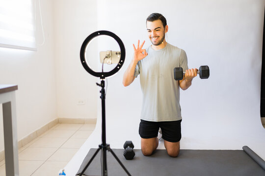 Young man giving online fitness class from home, holding dumbbell and making ok sign, recording virtual workout session