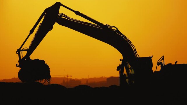 Excavator silhouette in golden sunset, digging and lifting earth, showcasing continuous action in a construction site with distant city skyline visible