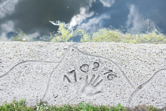 A human handprint with numbers carved into a stone pavement or concrete border next to a city park lake water.