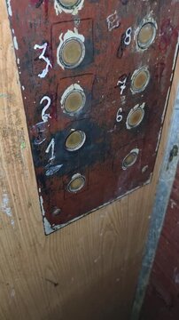 Close-up of a rusty and broken elevator control panel with round buttons and handwritten numbers. The white paint on the grimy surface evokes decay and abandonment.