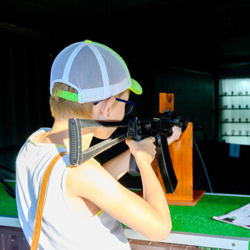 rear view of a boy in a baseball cap and glasses shooting from an automatic rifle on a wooden stand in a range