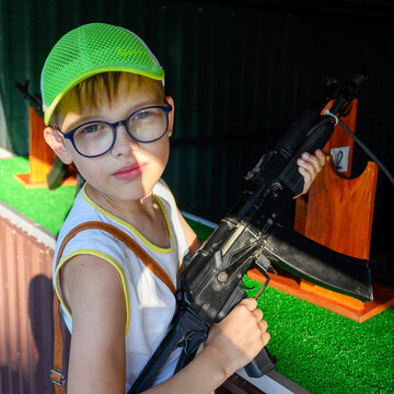 portrait of a boy in glasses and a bright green cap with a black automatic rifle in his hands near range stand