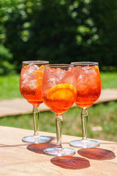 Three glasses of refreshing orange spritz cocktails with ice and citrus slices served outdoors on green sunny patio. Summer relaxation, outdoor dining, leisure, travel and social gathering concept.