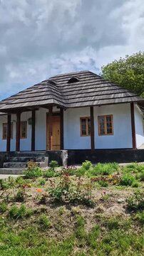 Traditional Moldovan peasant house with burned shingle roof. The cottage features white clay-plastered walls, wooden windows, and a porch supported by dark timber pillars, fronted by a blooming garden