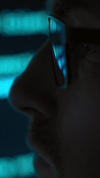 Side view close up, focused software engineer in eyeglasses looking at blue code running on screen in dark room, programming script reflects in his lenses. AI and data science concept. Vertical video