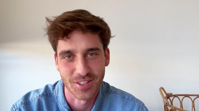 Casual young man with stubble and a denim shirt speaking directly to the camera during a video conference, expressing various emotions while having a pleasant conversation from home