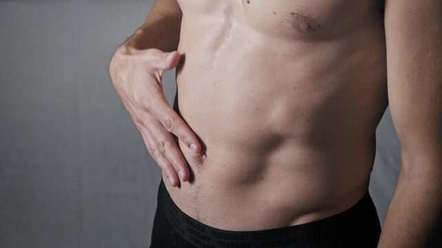 Close-up of a shirtless man with a lean muscular build touching his abdomen. He checks his defined abs and core muscles while feeling for potential physical discomfort.