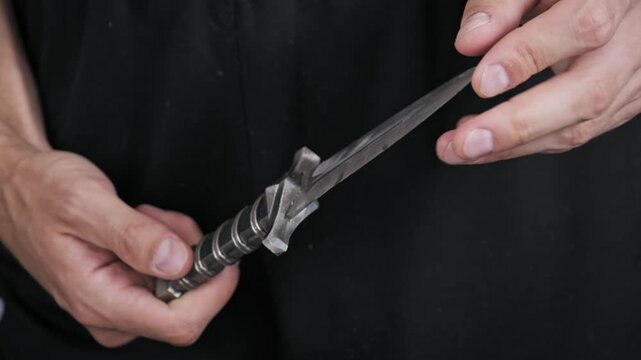Close-up of hands holding and carefully examining a small ornate dagger. The vintage weapon features a detailed hilt and a rough hand-forged metallic blade.