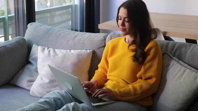Young adult woman sitting on the couch in a modern living room, working from home on her laptop computer, typing on the keyboard, and managing her online business with a focused expression
