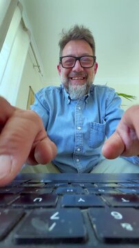 Low-angle perspective of a smiling middle-aged businessman working on his computer, with his fingers typing on the keyboard in the foreground and his happy face visible in the background