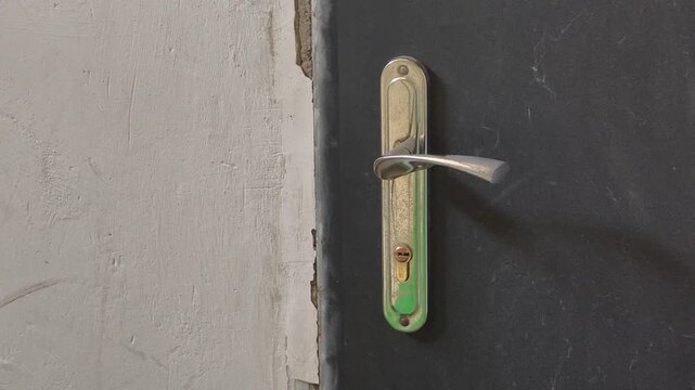 Close-up pan of a weathered silver door handle and lock on a dark textured door. The adjacent rough white wall adds to the old rustic and mysterious atmosphere of the entryway.