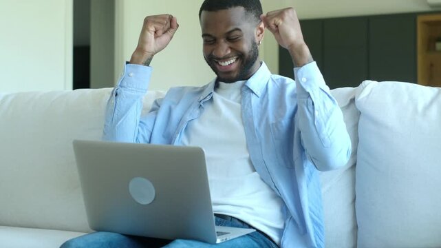 Excited african american man celebrating a win, reading good news on his laptop while sitting on the couch at home. Happy freelancer or student achieving success and feeling euphoric