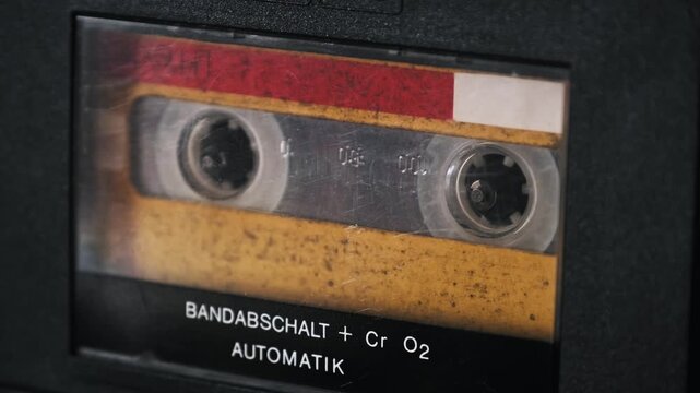 Macro shot of a vintage audio cassette playing in a retro tape deck with a spinning reel and scratched plastic. The worn red and yellow label creates a strong visual texture of 1980s music history.