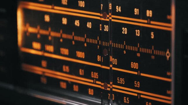 Macro of a vintage radio tuner needle moving across an illuminated orange frequency scale. Analog numbers and markings on the display highlight the search for broadcast stations.