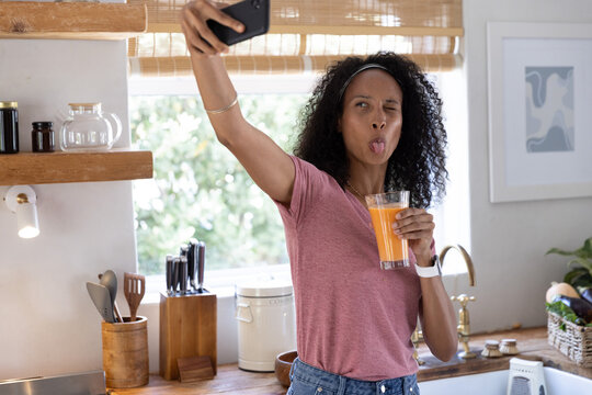Adult woman is raising smartphone and glass of orange juice and making playful face in kitchen