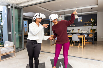 Obraz premium Diverse female friends holding hands and using white VR headsets on black mats near glass doors
