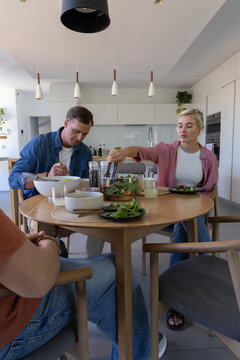Three adults sitting around round wooden dining table in modern kitchen serving salad with tongs