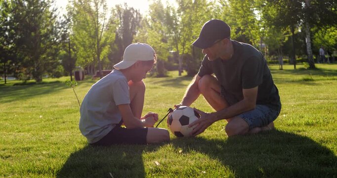 Father and son preparing soccer ball with hand pump while sitting on green grass in park at sunset. Dad checks air pressure as boy pumps ball. Teamwork, cooperation and family interaction. Vertical