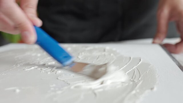 Hand applies white textured paste on canvas with a small spatula creating a relief surface. Concept of DIY textured art and handmade wall decor.