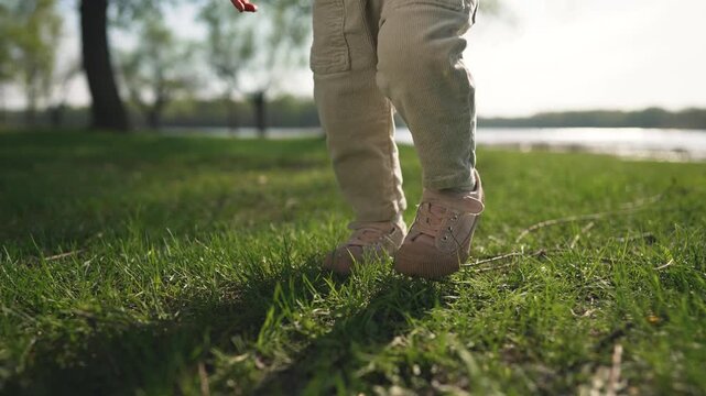 Toddler takes first steps outside father hand guiding. Father supports toddler early steps fresh air. Toddler takes his first steps with his father on outside. First steps toddler and father outside.