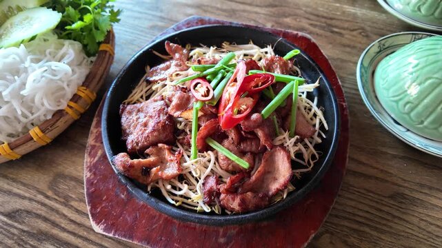 A close-up of a black bowl filled with crispy pork slices, bean sprouts, and sliced red chili peppers garnished with green onions on a wooden table in Hoi An, Vietnam