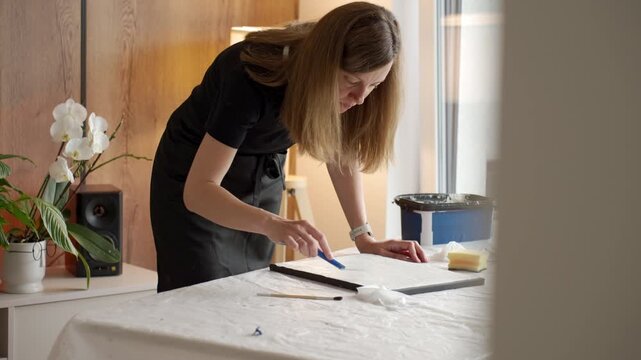 Female artist creating a textured picture on canvas applying white plaster paste with a blue palette knife at table. Concept of handmade painting and creative hobby
