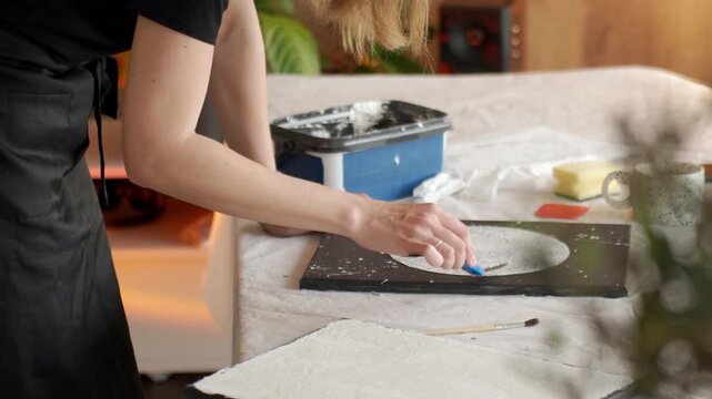 Woman spreads white plaster paste on black canvas with palette knife at covered table. Concept of DIY abstract art handmade decor and creative process.