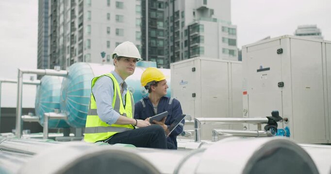 Two industrial engineers collaborate on system diagnostics, using a laptop and tablet to inspect a large HVAC installation on a building rooftop.