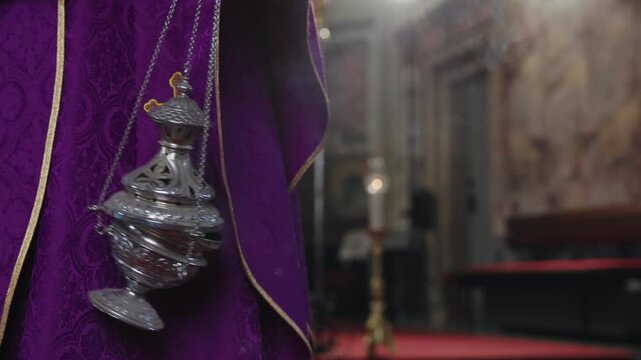 Priest in a purple clergy robe, swinging an incense burner, a thurible releasing fragrant smoke during a mass at a Catholic church.
