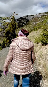 Vertical view of woman wearing puffer jacket walking on a hiking path by a mountain
