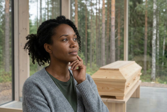 Pensive Black woman reflecting on a natural wooden coffin in a modern forest chapel. Eco-friendly funeral and burial planning concept. Mourning and remembrance in a woodland setting