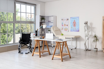 Fototapeta premium Modern physical therapy office interior with wheelchair, examination couch, and anatomical charts for rehabilitation