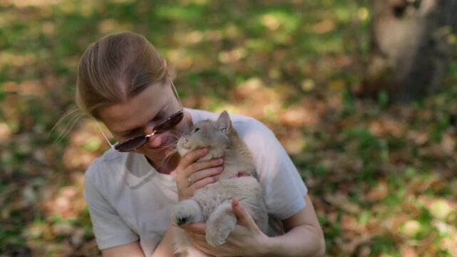 Woman holds cat gently while sitting under trees shade. She hugs her pet tenderly. Cat looks calm, eyes half-closed in trust. Sunlight filters through leaves, soft and warm.